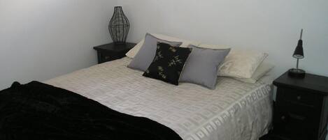 Una cama bien hecha con una manta negra, una colcha estampada y varios almohadones. Hay una mesita de noche con una lámpara y un adorno en la pared.