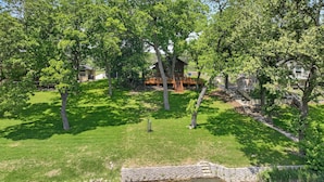 Un espace verdoyant bien entretenu, agrémenté d’une terrasse en bois et d’un muret en pierre, le tout entouré d’arbres.