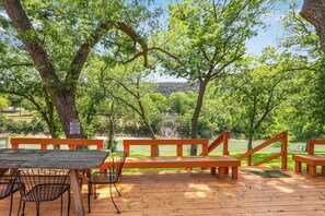 Een terras buiten met houten banken en tafels, omringd door bomen en uitzicht op een meer.