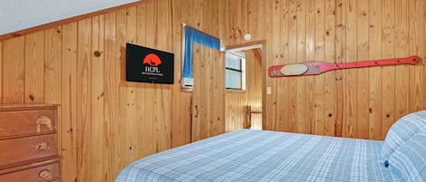 A bedroom with wooden walls, a bed with a plaid bedspread, a dresser, a sign with a logo, and a paddle on the wall.