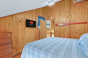A bedroom with wooden walls, a bed with a plaid bedspread, a dresser, a sign with a logo, and a paddle on the wall. - Perfect Spot on the Guadalupe River! Sleep up to 10 and Float all day! (New Braunfels)