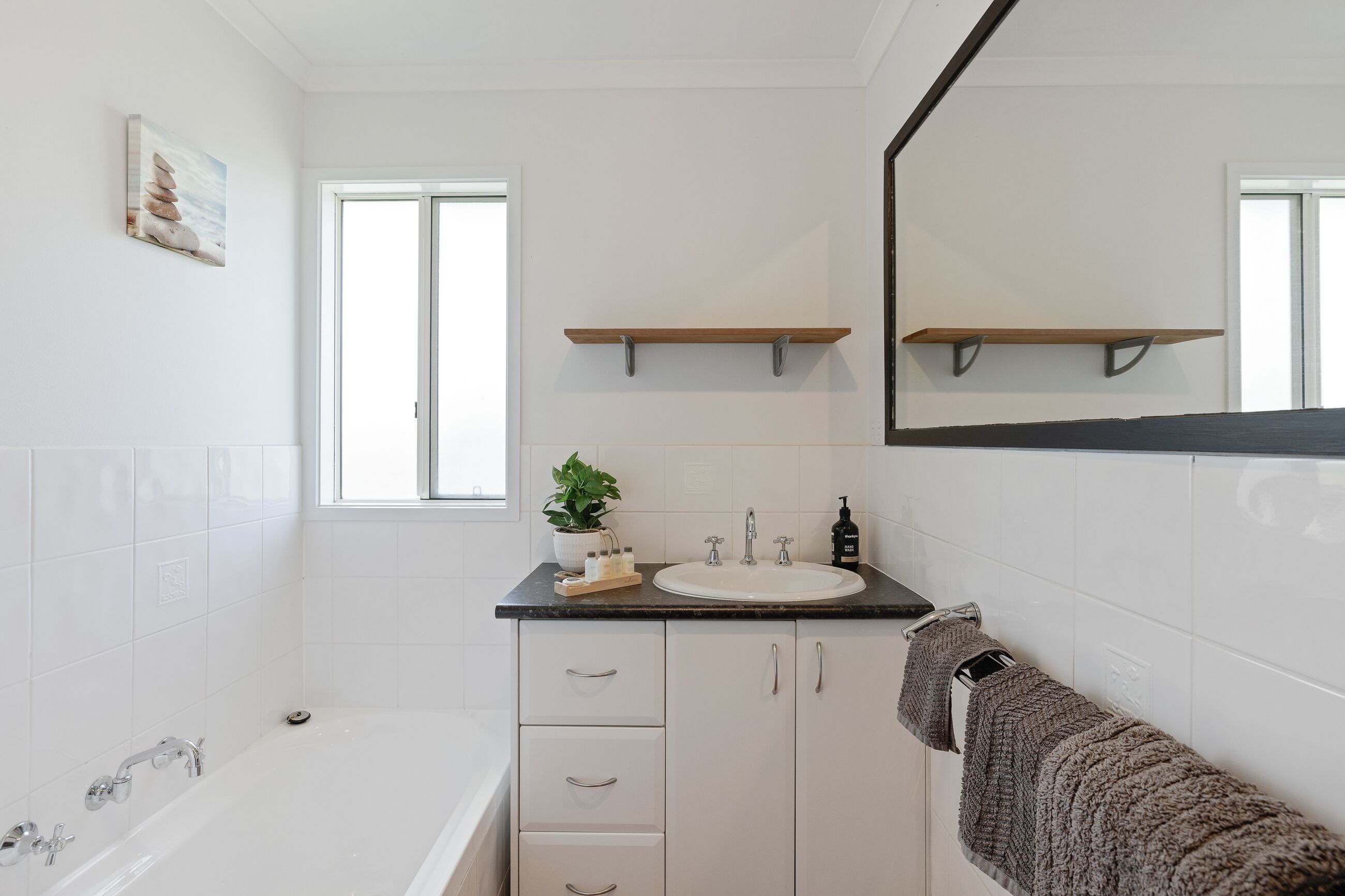 A modern bathroom with a bathtub, white cabinetry, a sink with a mirror above, and a window.