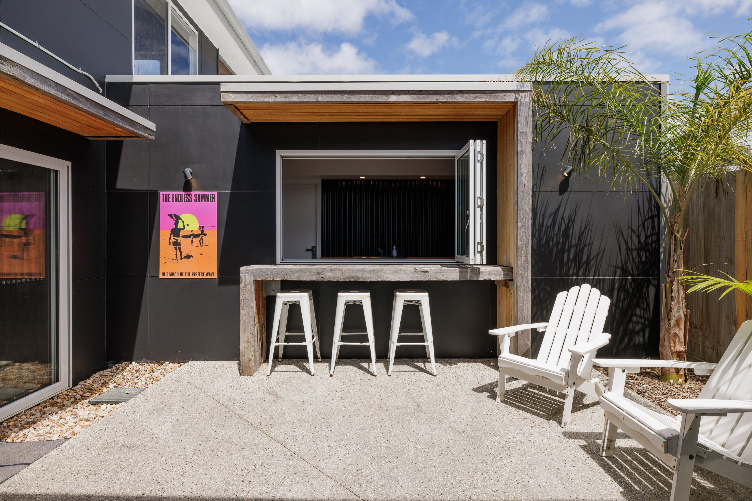 Un espace de détente extérieur moderne, doté d’un sol en béton, de trois tabourets de bar blancs et de deux fauteuils Adirondack blancs.