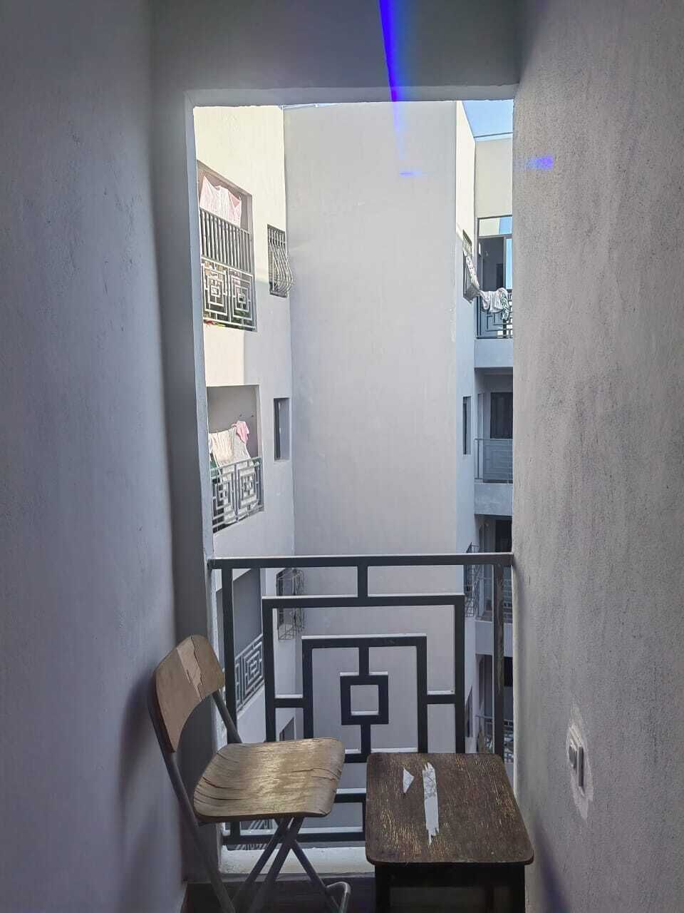 Un balcon avec une chaise et une petite table, une vue sur les immeubles voisins et un luminaire bleu.