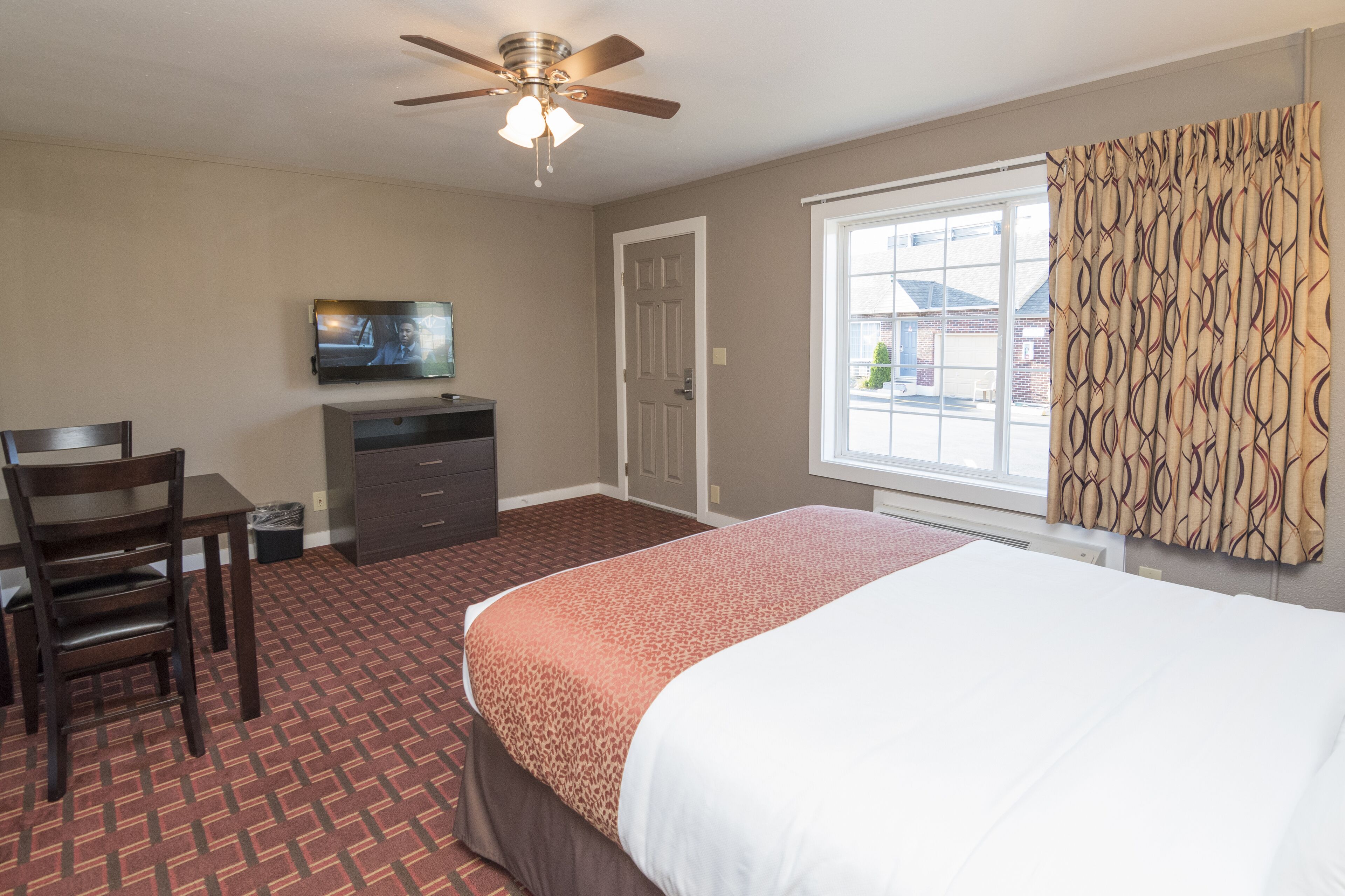 Quarto de hotel com cama, escrivaninha, cadeira, televisão e janela com cortinas.