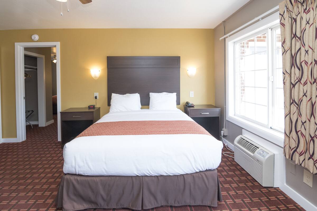 A hotel room with a large bed, two bedside tables, a heater, a window with curtains, and a door leading to a closet.