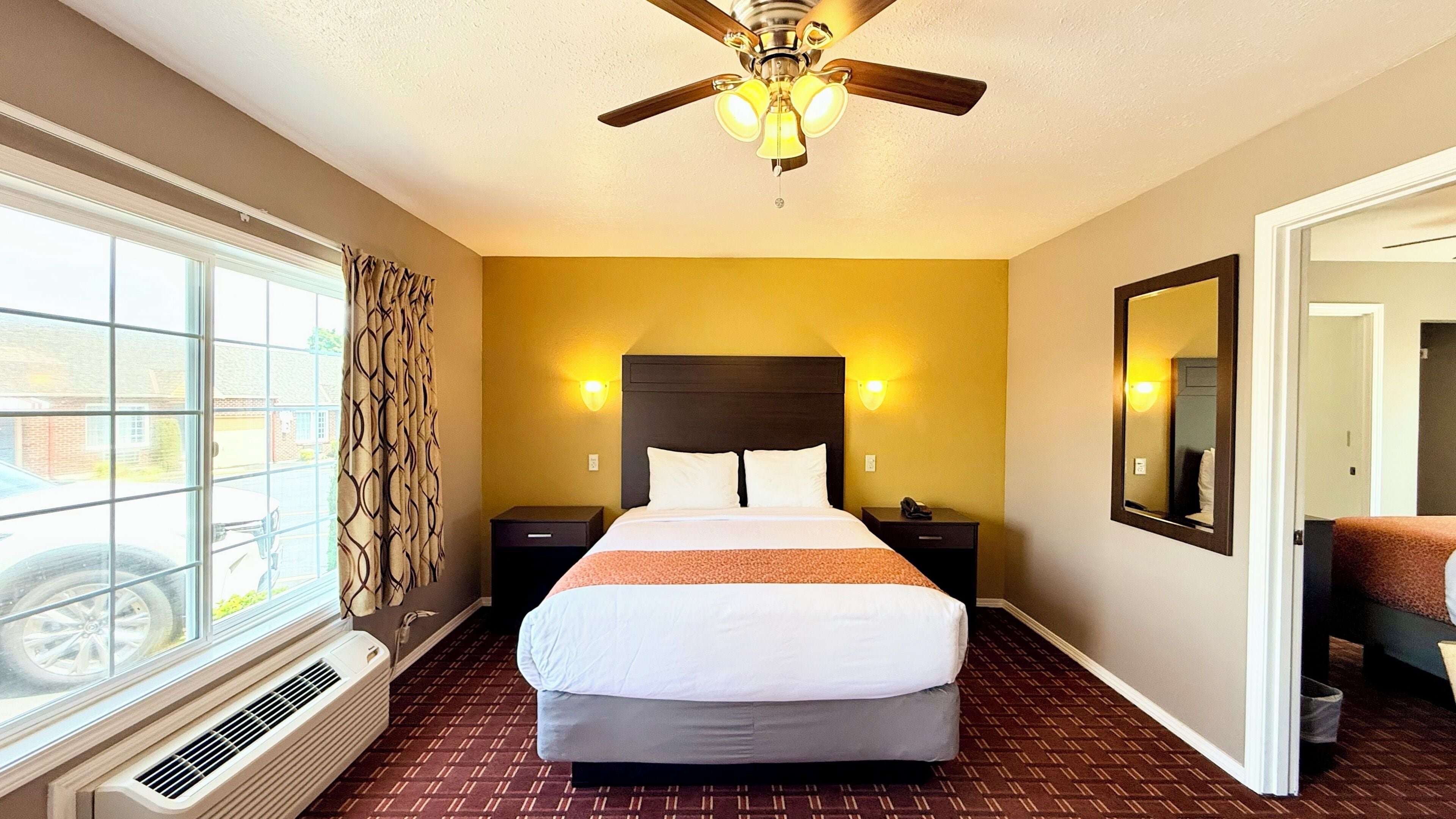 Une chambre d’hôtel avec un lit, un ventilateur de plafond, une fenêtre avec des rideaux, un miroir fixé au mur et une porte.