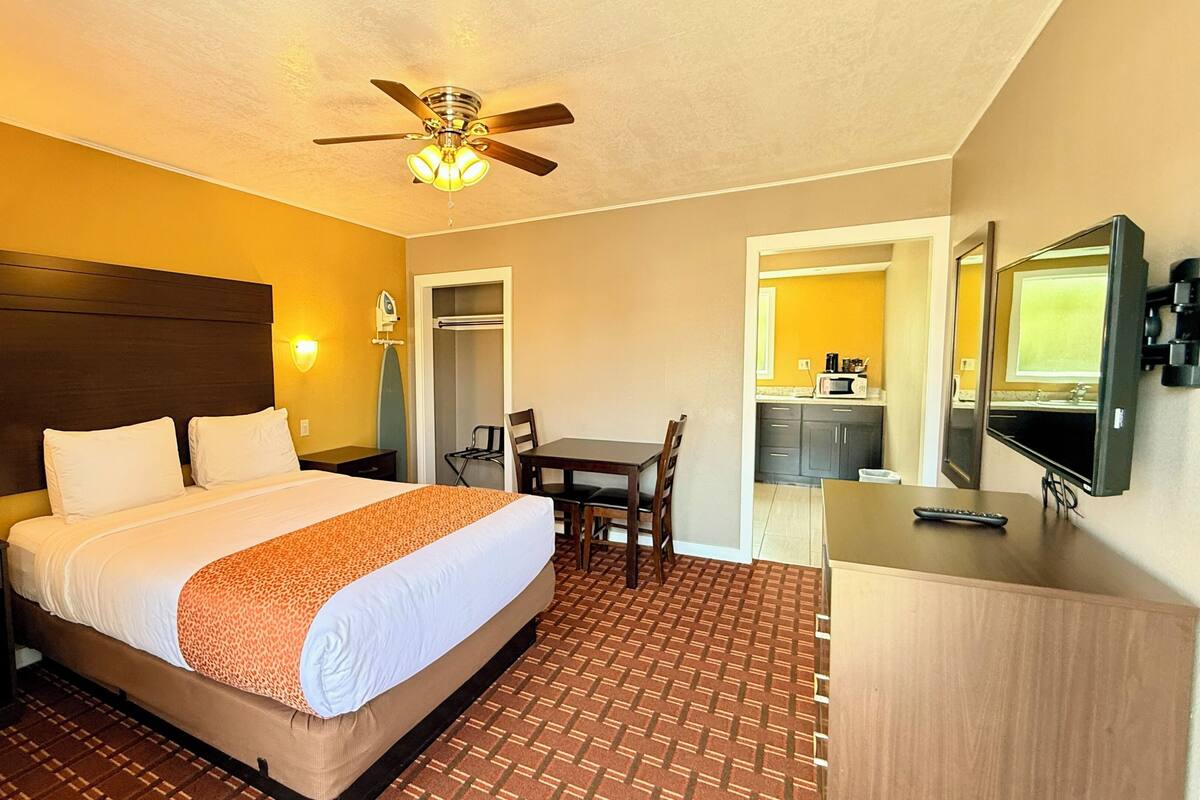 A hotel room with a bed, a desk, a chair, a wardrobe, a mirror, a ceiling fan, and a television.