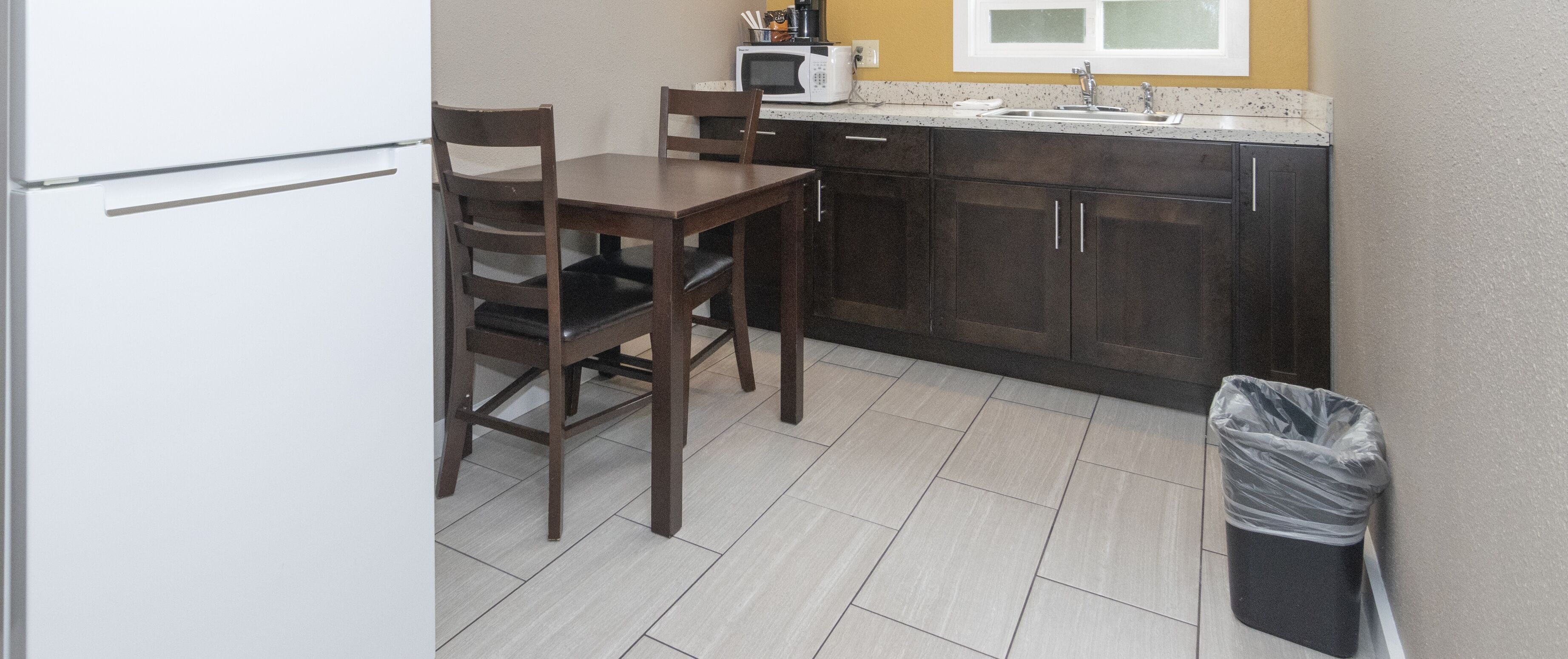 A compact kitchen with dark wooden cabinets, a white countertop, a microwave, and a trash bin.