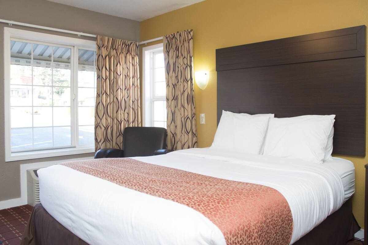 A hotel room with a large bed, a chair, a window with curtains, and a wall-mounted light.