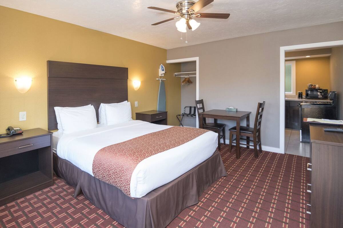 A hotel room with a bed, bedside table, lamp, and a kitchenette area.