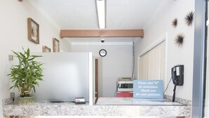 A reception area with a marble counter, a welcome mat, a potted plant, and a sign offering assistance. - Capitol Hill Motel (Portland)