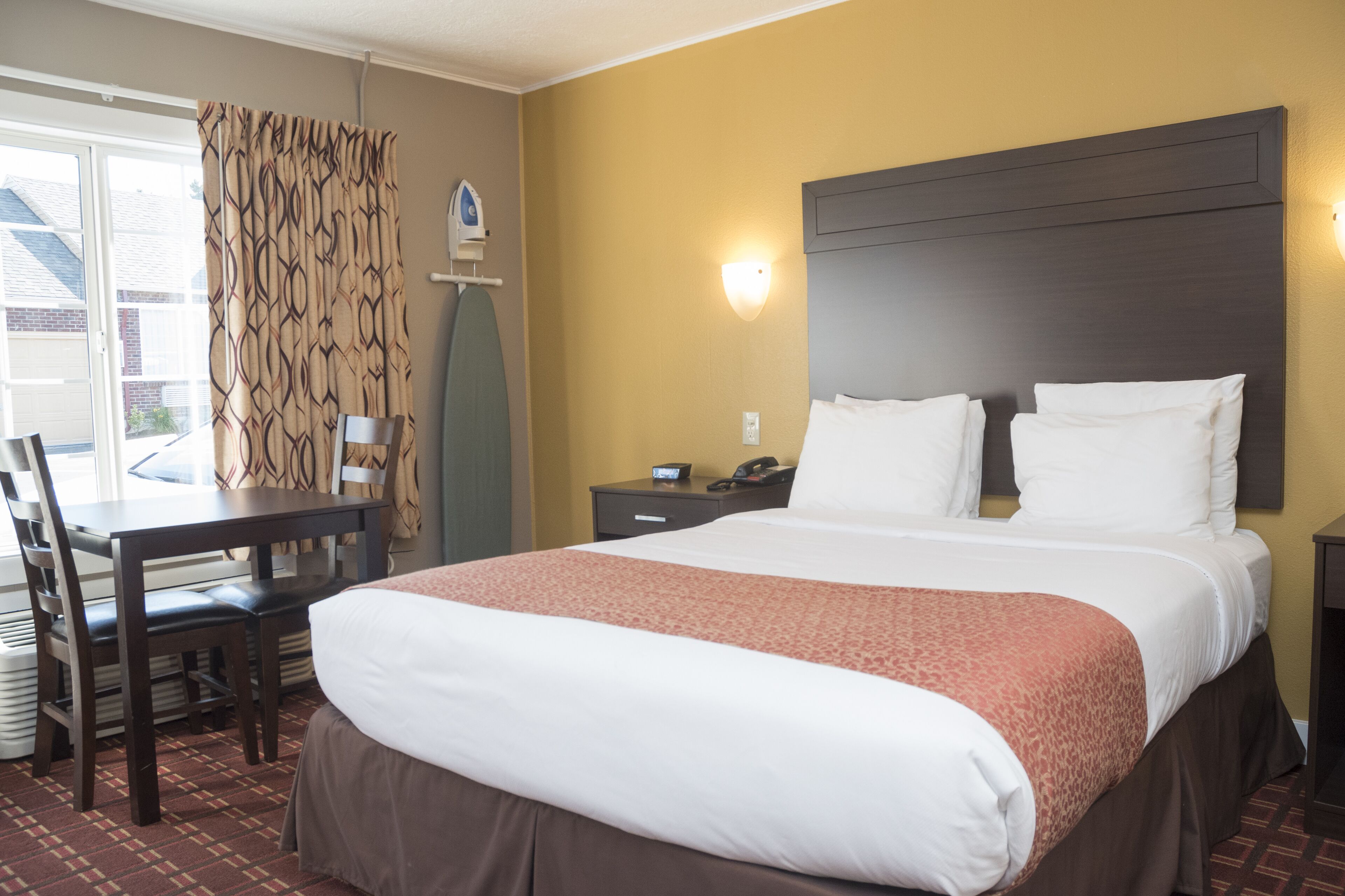 A hotel room with a large bed, a desk with a chair, a nightstand, and a window with curtains.