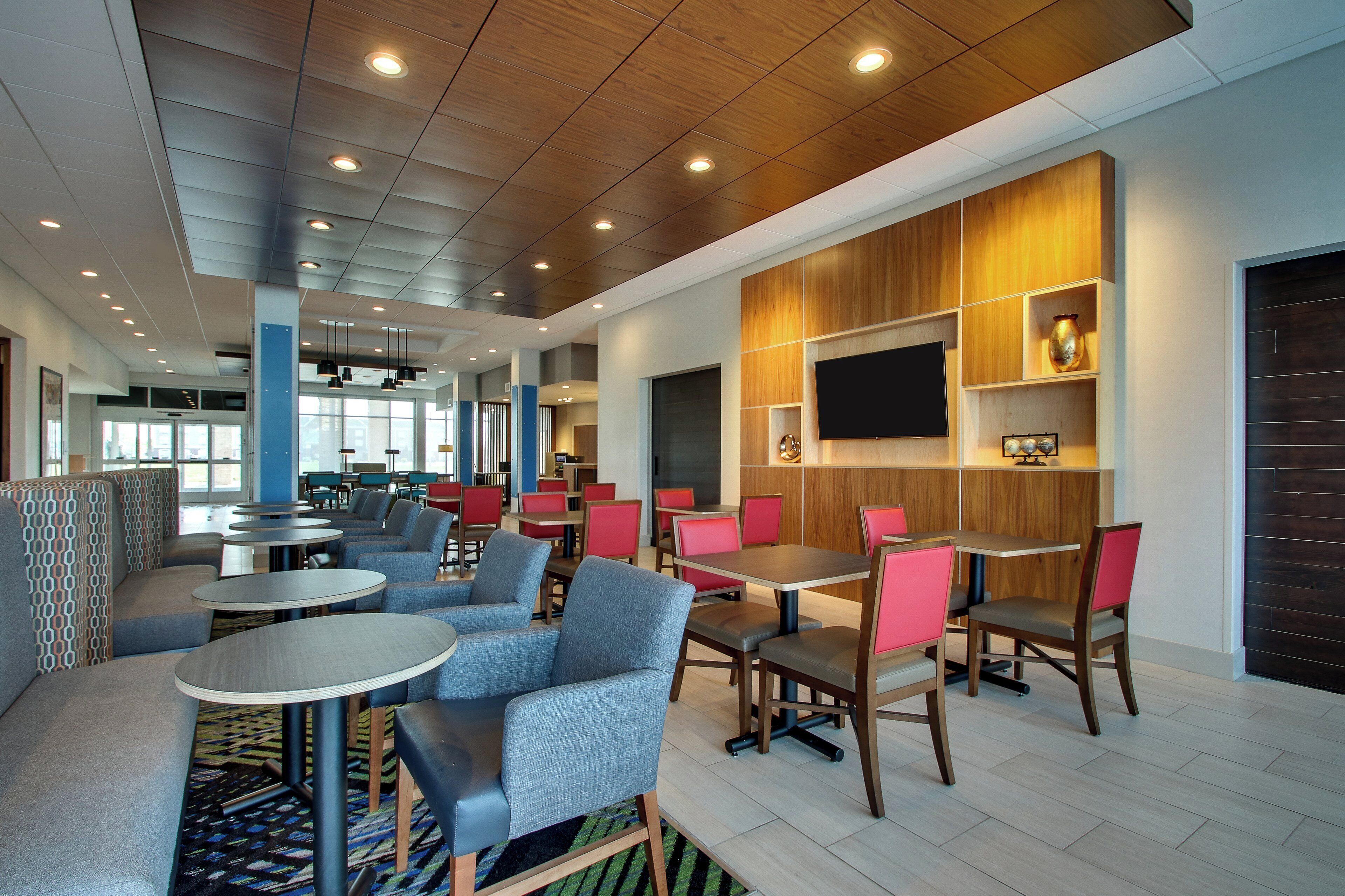 A modern hotel lobby with seating areas, a flat-screen TV mounted on a wooden panel, and a patterned carpet.