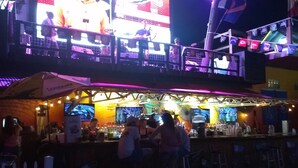 A bar with a large screen displaying a performer. There are people seated at the bar and a white picket fence in the foreground. - Gipson Inn Motel (Fort Walton Beach)