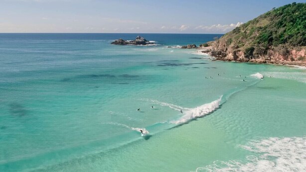 Une plage côtière aux eaux turquoise et limpides, bordée de rochers et surplombée d’une colline verdoyante.