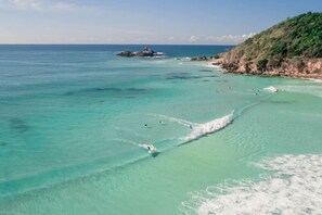 Een kuststrand met helder turquoise water, rotsformaties en een heuvelachtig achtergrondlandschap.