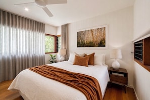 Een slaapkamer met een groot bed, een plafondventilator, een raam met gordijnen, een schilderij aan de muur en een nachtkastje met een lamp.