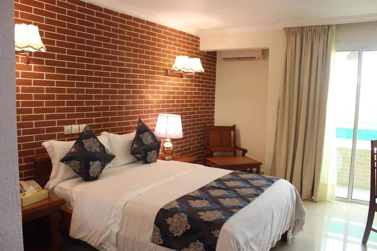 A hotel room with a brick wall, a bed with white bedding and patterned pillows, a nightstand with a lamp, and a chair near a sliding glass door.