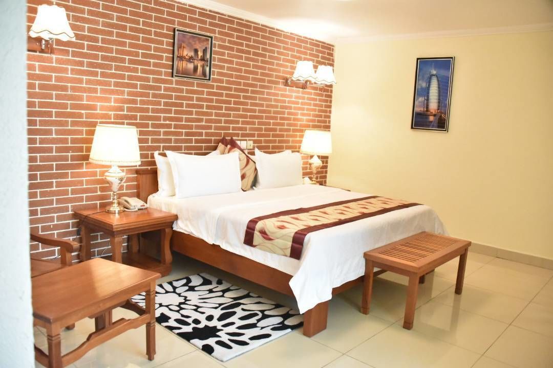 Une chambre d’hôtel avec un mur en briques, un lit avec une literie blanche, deux lampes de chevet, un banc en bois, une petite table et un tapis à motifs.