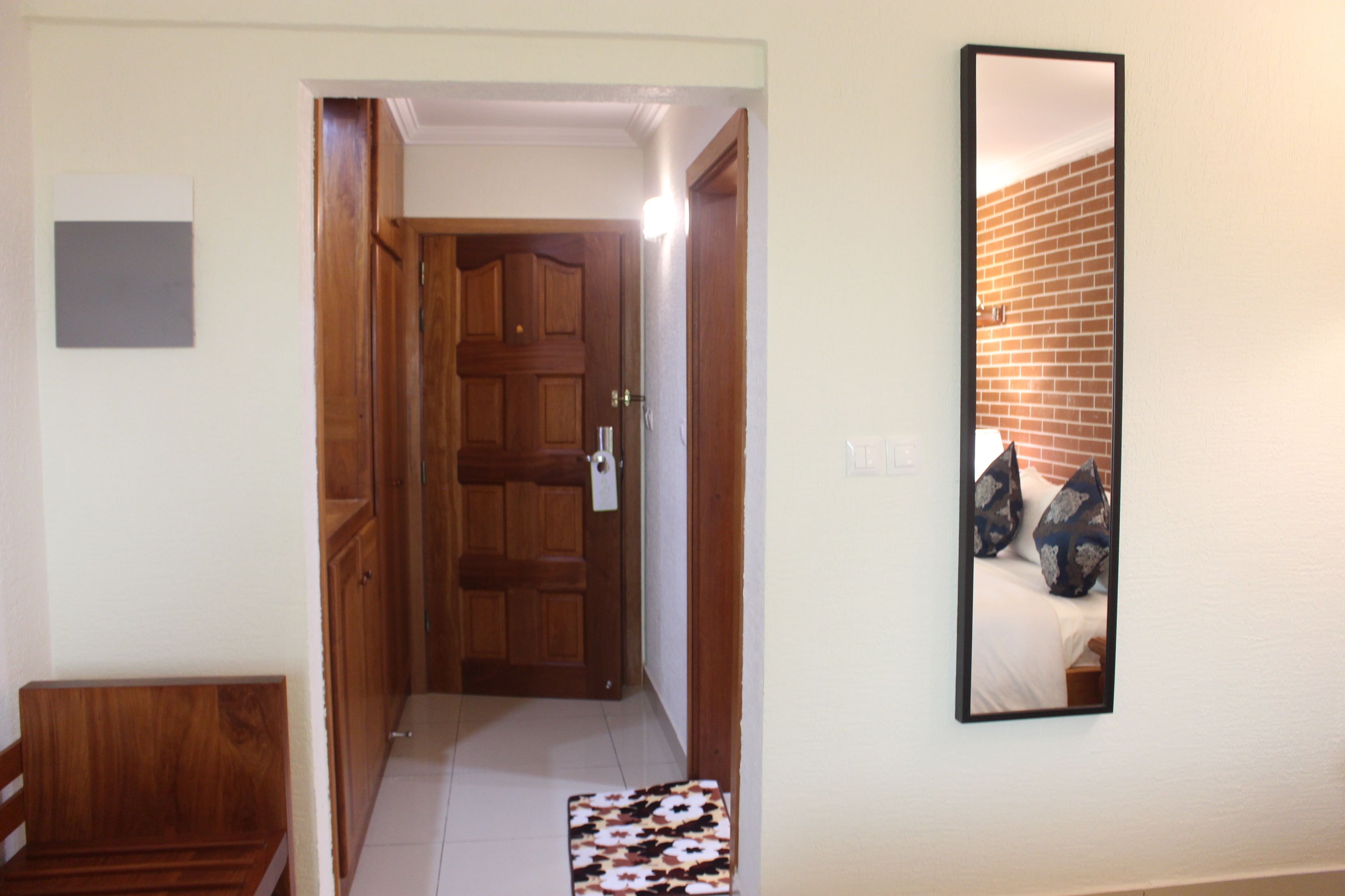 A hallway with a wooden door, a small rug, and a mirror reflecting a bed with pillows.