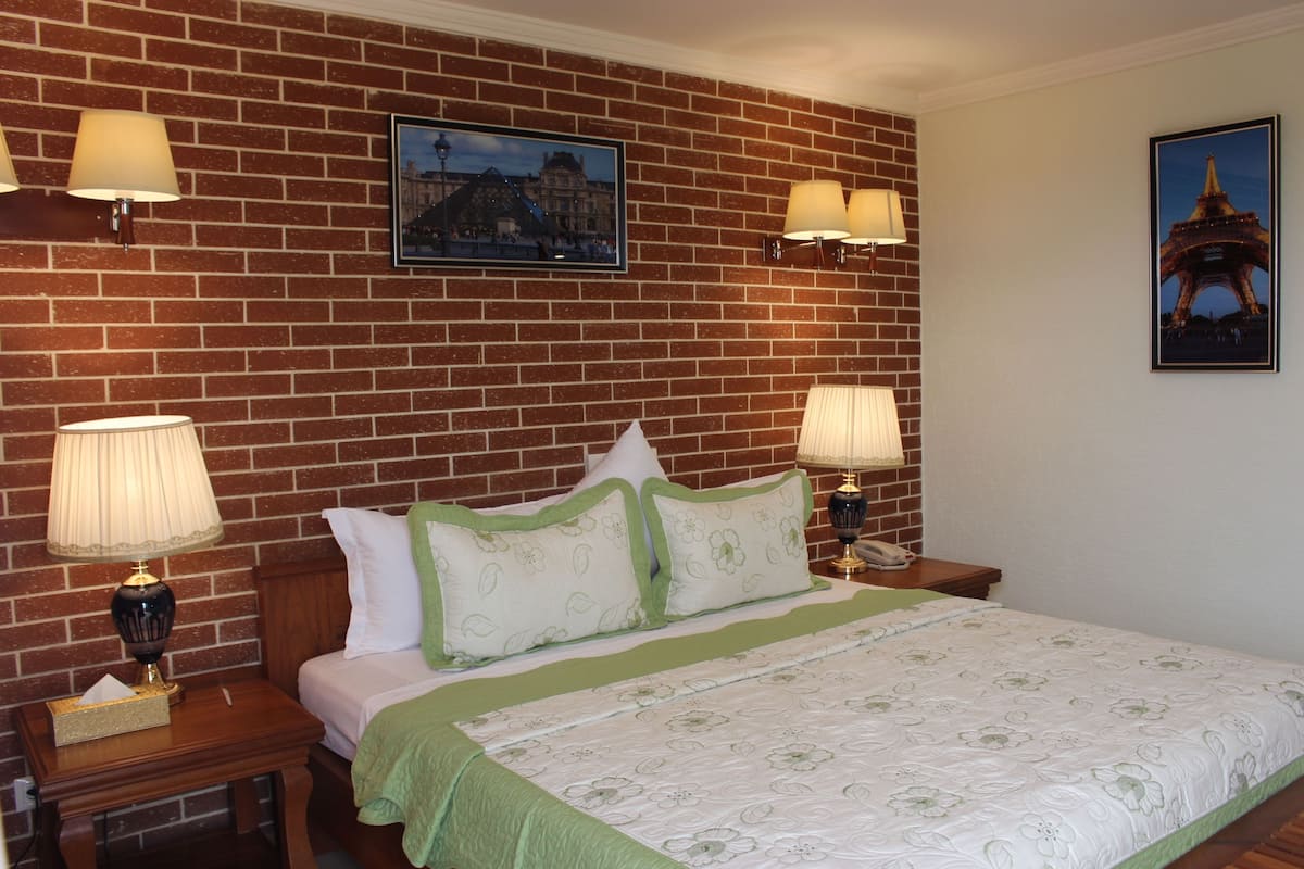 A hotel room with a brick-patterned wall, a bed with green and white bedding, two bedside lamps, and two framed pictures on the wall.