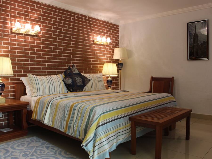 A bedroom with a brick wall, a bed with striped bedding, two bedside lamps, a wooden bench, and a framed picture on the wall.