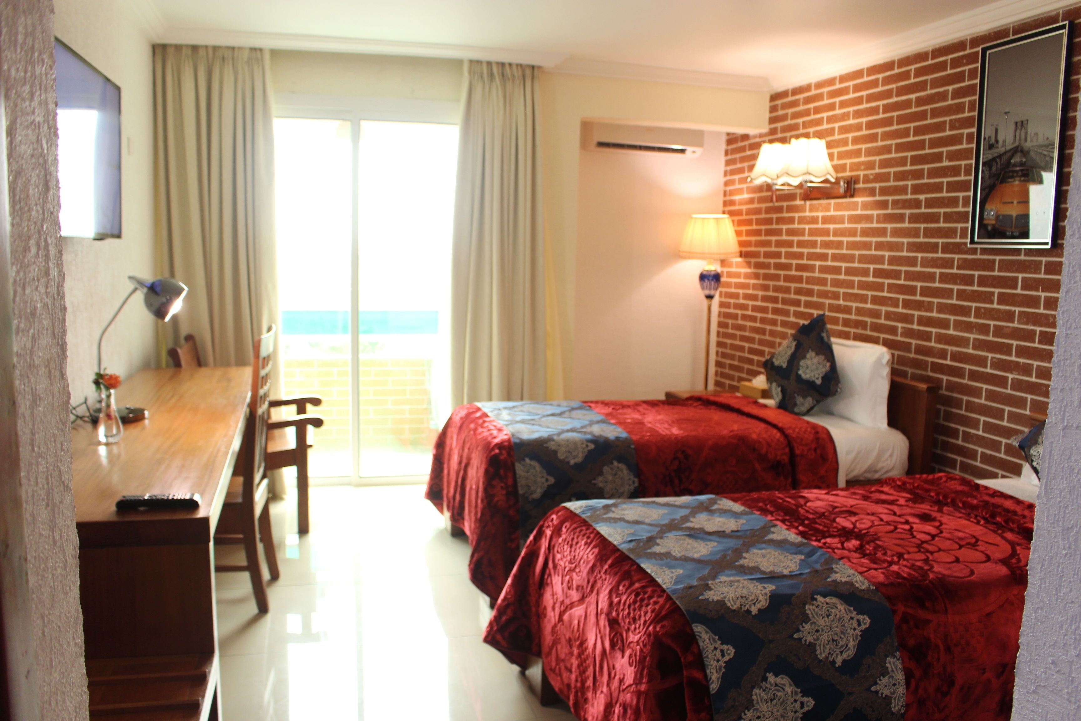 A hotel room with two beds, a desk, and a chair. There is a window with curtains, a lamp, and a picture on the wall.