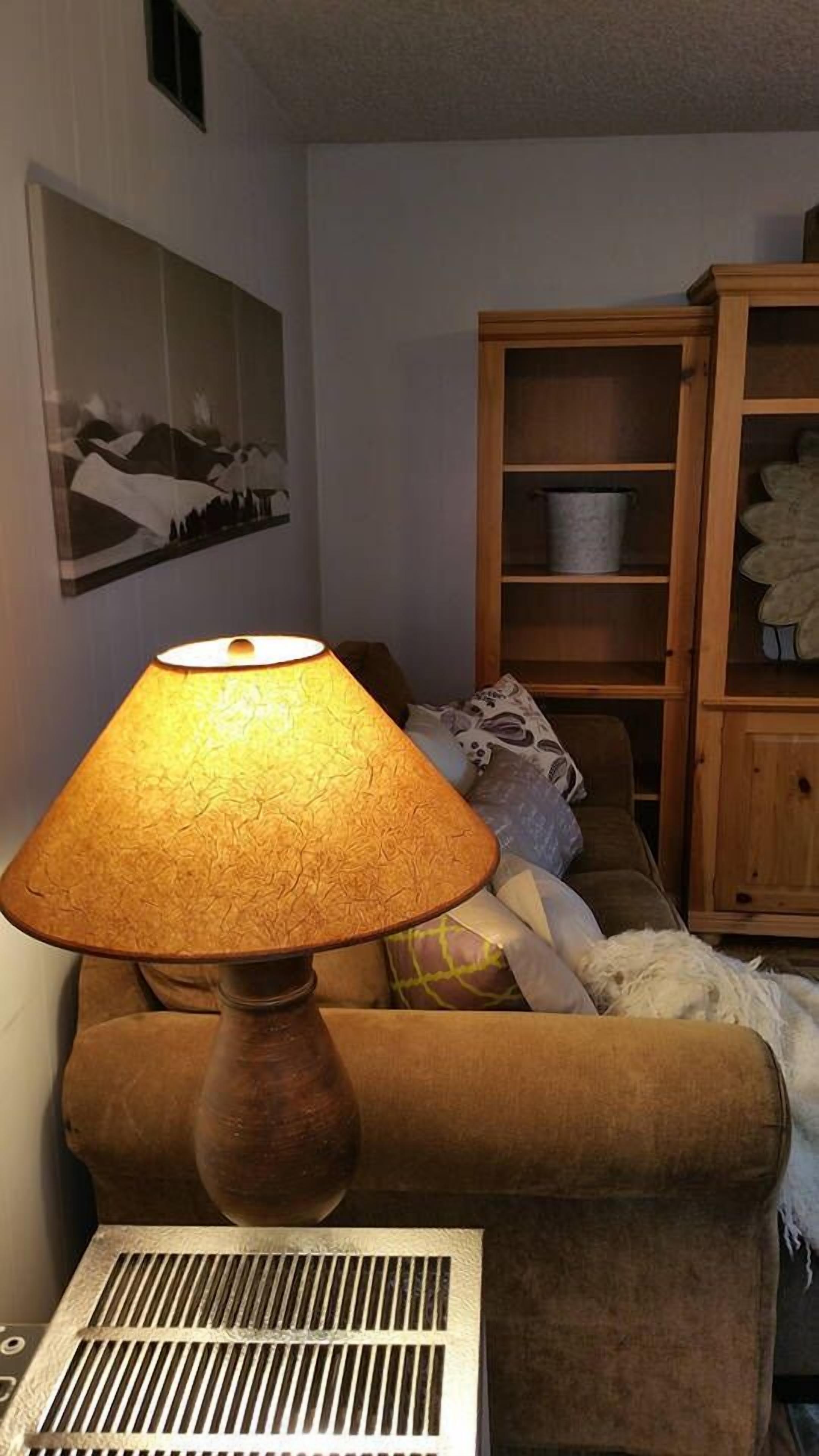 Una habitación con una lámpara sobre una mesa, un sofá, un radiador y un aparador de madera con estantes.