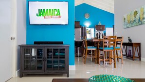 A living room with a TV displaying 'Jamaica Travel Channel', a wooden cabinet, a dining area with chairs, and a decorative plate on a table. - Casa Princess Ocho Rios Vacation Villa (Ocho Rios)