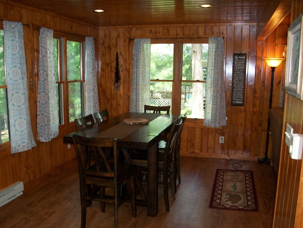 Une salle à manger en bois avec une table et des chaises, de grandes fenêtres ornées de rideaux à motifs, un panneau mural et un lampadaire.