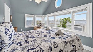 A bedroom with a large bed, a ceiling fan, and multiple windows offering a view of the sea and a pier.