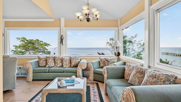 A spacious living room with two green sofas, a coffee table, and a chandelier. Large windows offer a view of the sea and a pier.