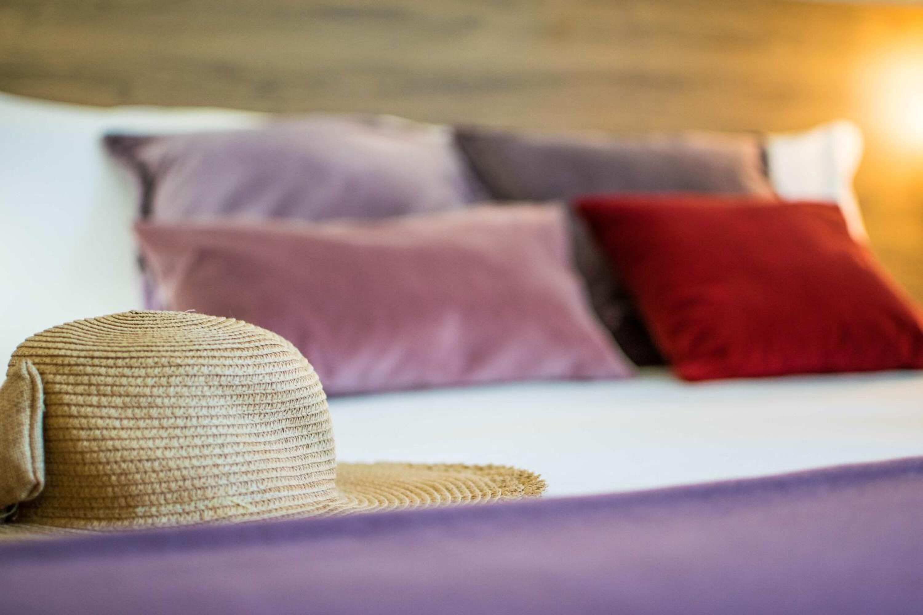 A bed with a straw hat on it, and pillows in shades of purple and red.
