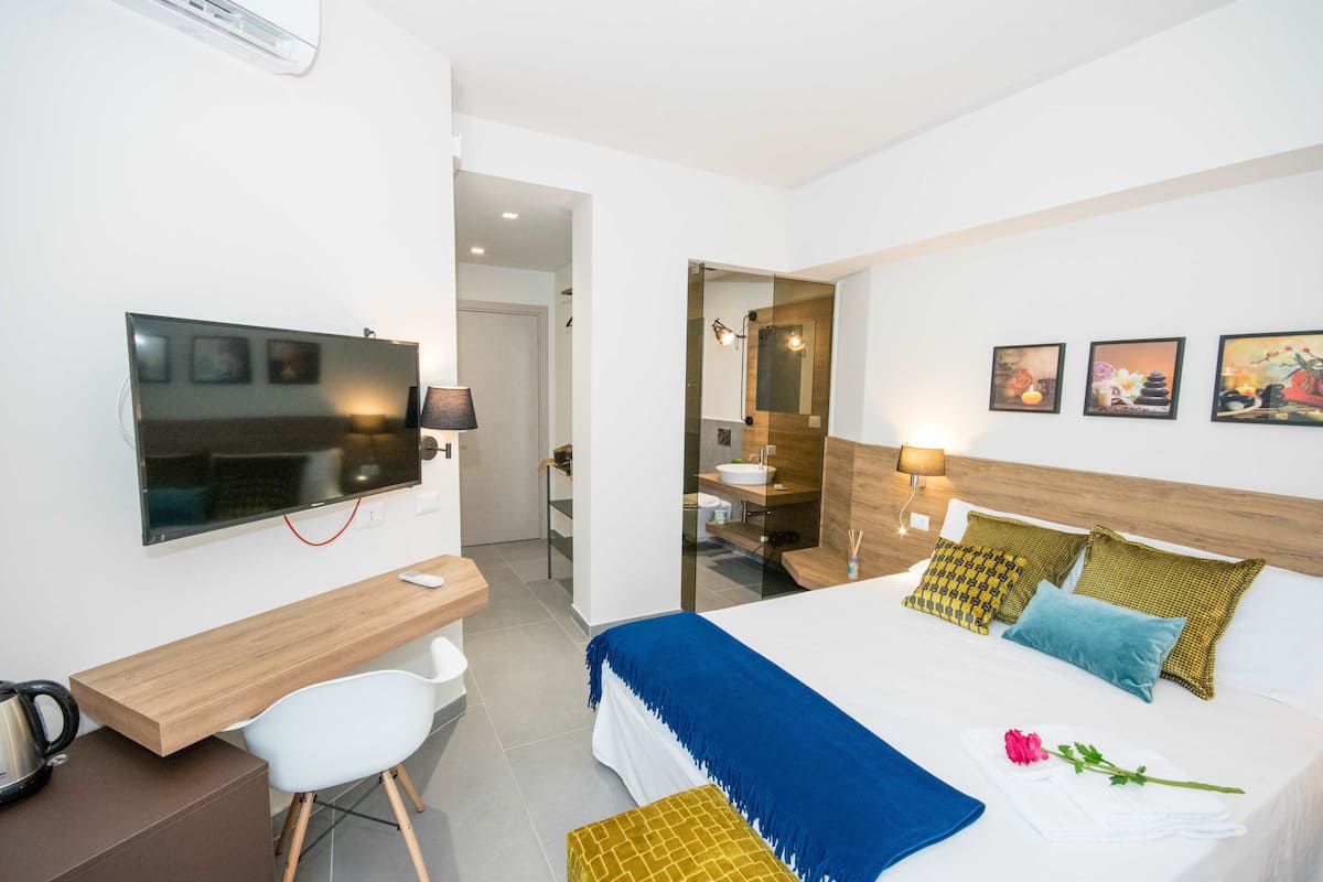 A modern hotel room with a bed, bedside table, chair, desk, TV, and a bathroom visible through an open door.