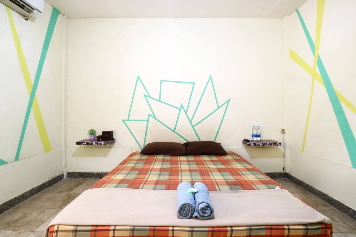 A hotel room with a bed, pillows, a blanket, and towels. There are two side tables with bottles and a small plant.