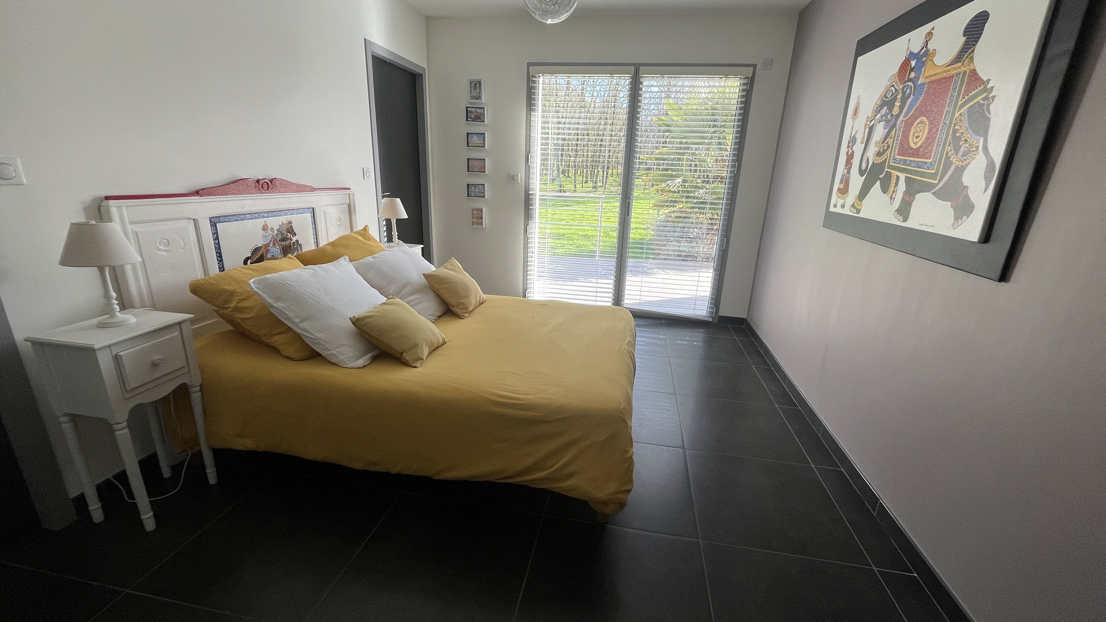 Une chambre à coucher comprenant un lit, une table de chevet, une lampe, une fenêtre avec des stores et un tableau encadré accroché au mur.