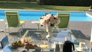 A glass table with breakfast items, a vase of flowers, and chairs by a pool. - Domaine Georges V (Saint-Junien)