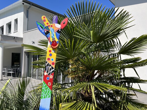 Une sculpture colorée représentant une girafe, placée devant un bâtiment à la façade blanche, bordé de palmiers.