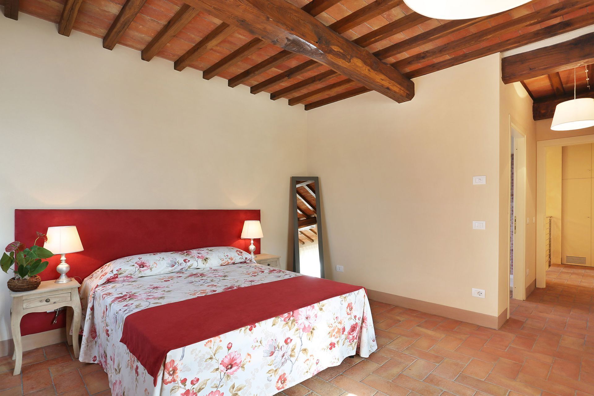 Camera da letto con testiera rossa, copripiumino floreale, due abat-jour, uno specchio e pavimento piastrellato.