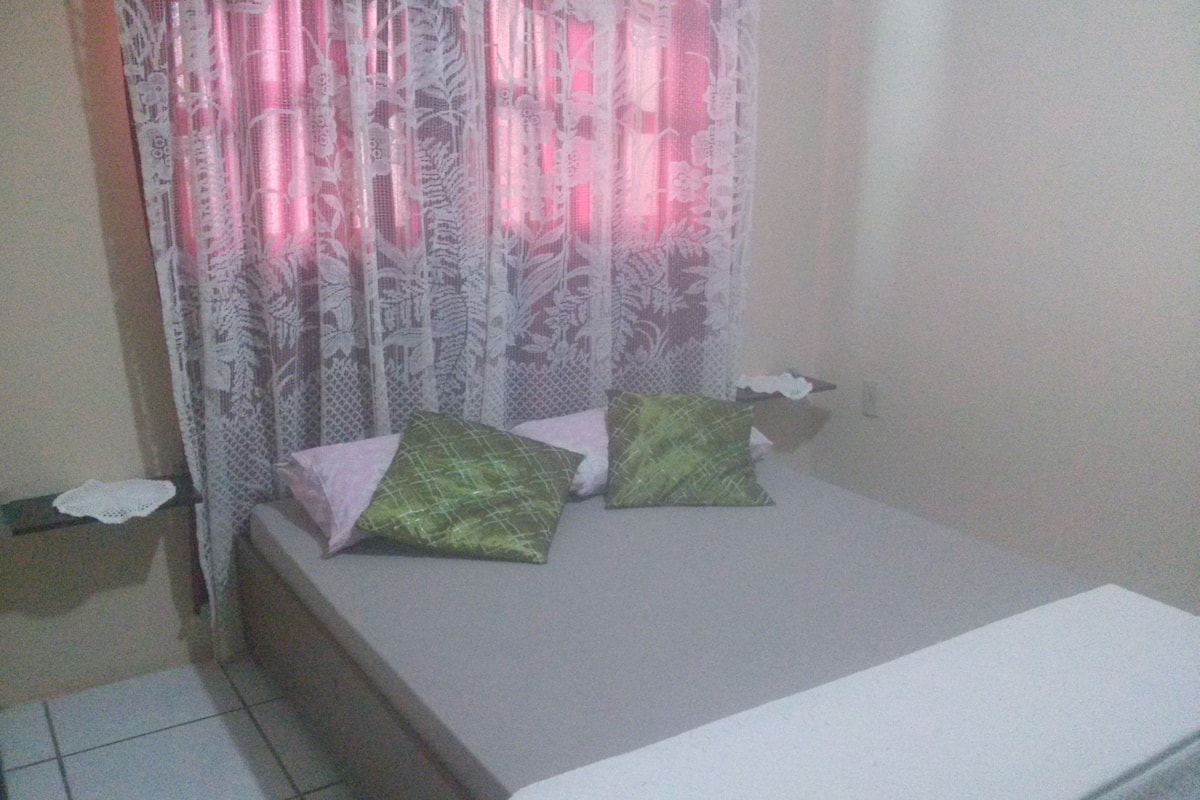 A small, simple room with a bed, two pillows, a lace curtain, and a tiled floor.