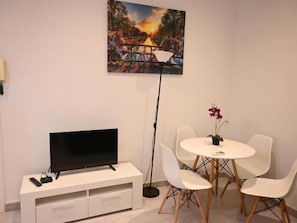 Une pièce avec un téléviseur posé sur un meuble blanc, une table ronde entourée de chaises et un tableau accroché au mur.