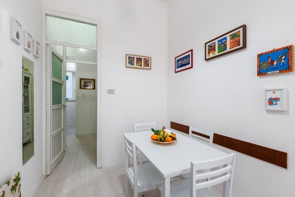 Un coin repas avec une table et des chaises blanches, un saladier de fruits posé sur la table, et divers tableaux encadrés accrochés aux murs.