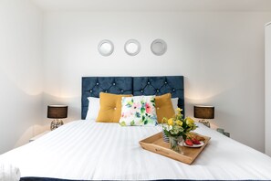 A neatly made bed with a blue headboard, white bedding, and a floral pillow. There are two bedside lamps, a tray with flowers and food, and three wall-mounted mirrors. - KVM - City Apartments (Peterborough)