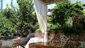 Un espacio al aire libre con sillas de mimbre y una mesa con cubierta de vidrio, rodeado de abundante follaje y un muro de piedra.