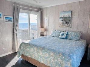 A bedroom with a large bed, a view of the sea through a sliding door, and a painting of reeds on the wall.