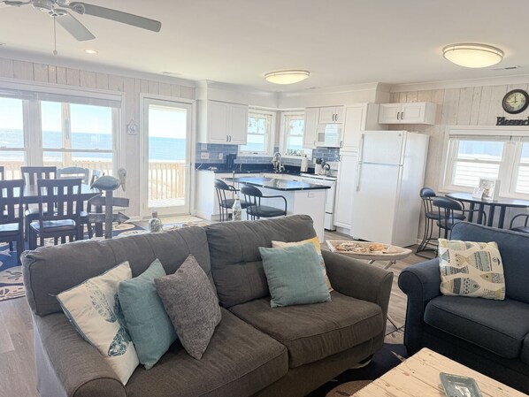 A living room with a gray sofa, a dining area with a table and chairs, a kitchen with white cabinets, and a view of the ocean through large windows.