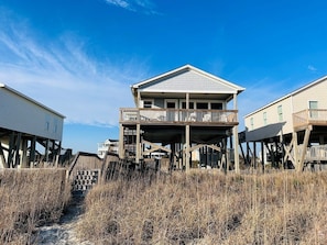 A beachfront house with a balcony, wooden stilts, and a sandy path leading to it.