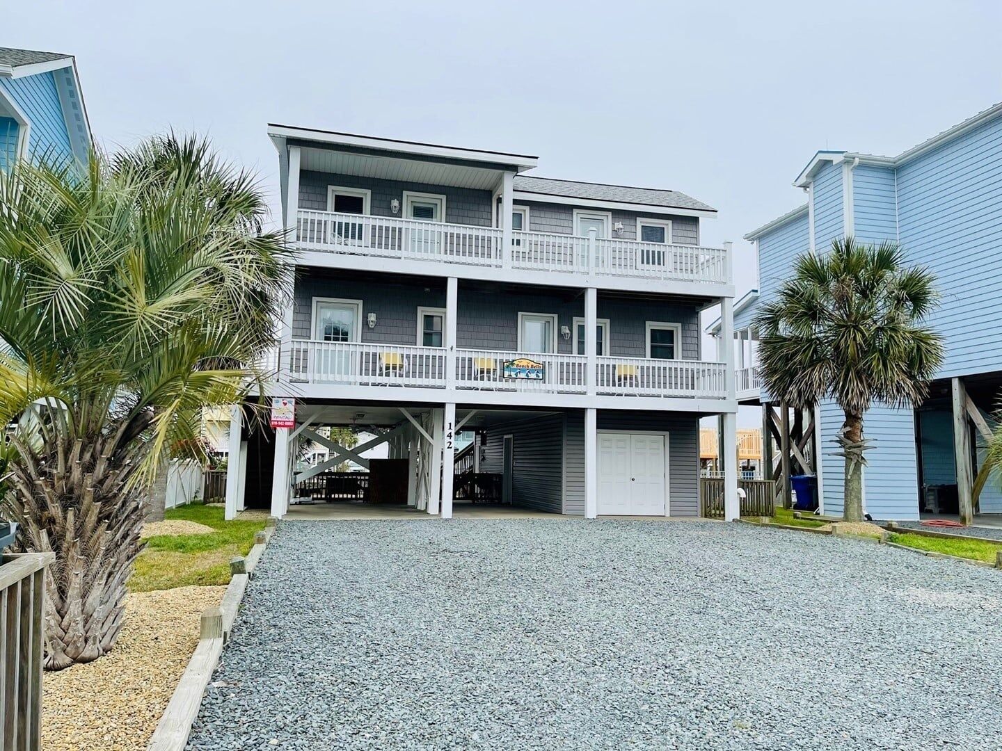 A multi-story building with a balcony, a sign indicating a beach resort, and a gravel driveway.