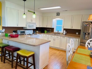 A kitchen with white cabinets, a microwave, a stove, and a sink. There are stools and a table with placemats. - Beach Bells 4 Bedroom Home by RedAwning (Supply)